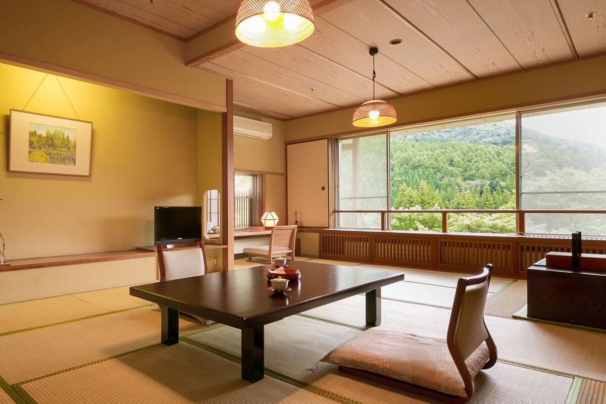 Una habitación japonesa tradicional con suelo de tatami, una mesa baja, un televisor y un ventanal con vistas a un bosque.