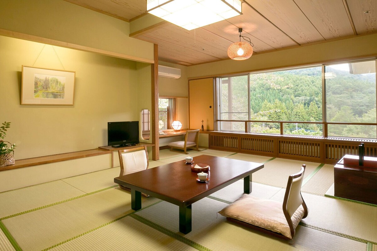 A traditional Japanese room with tatami flooring, a low table, a television, and a large window with a forest view.