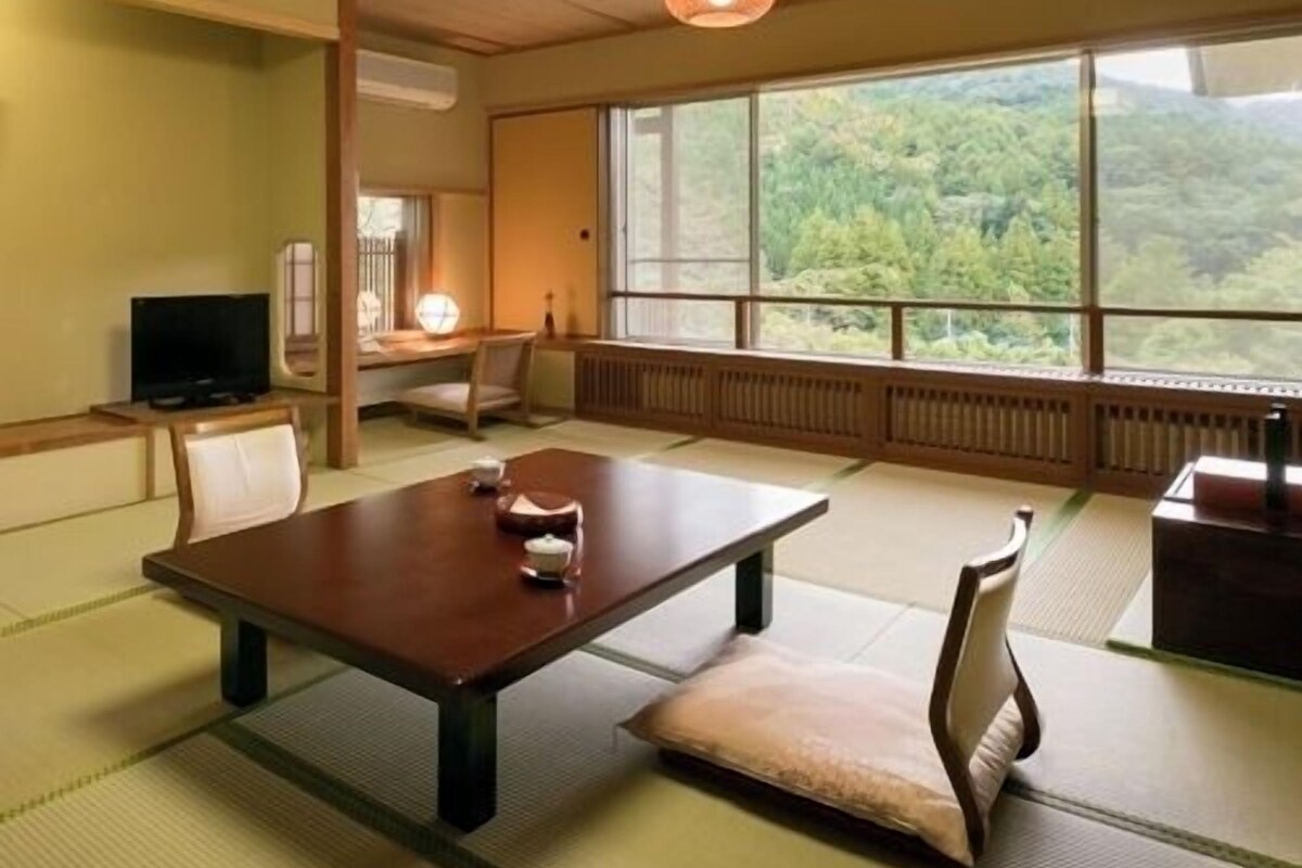 A traditional Japanese room with tatami flooring, a low table, a television, and a large window with a view of greenery.