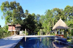 Una piscina con una estructura de techo de paja, rodeada de abundante vegetación y un edificio con fachada roja.