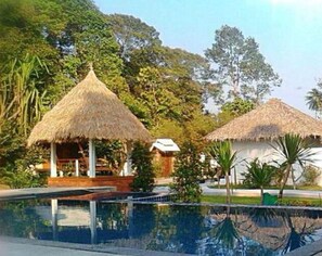 A pool area with thatched-roof structures, surrounded by lush greenery. - Hallo Villa (Khanom)