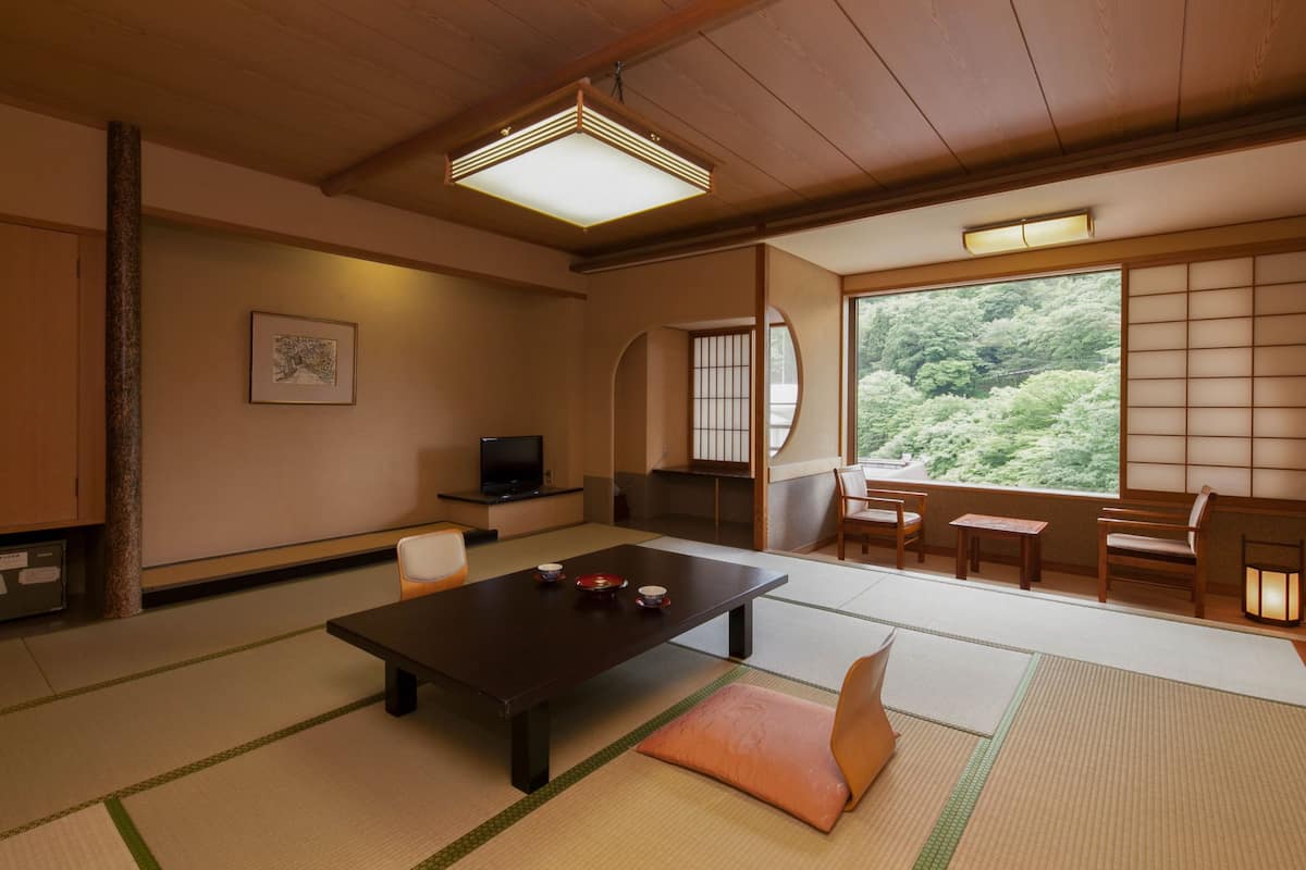 A traditional Japanese room with tatami flooring, a low table with a cushion, a television, and a view of a forest through a shoji screen.