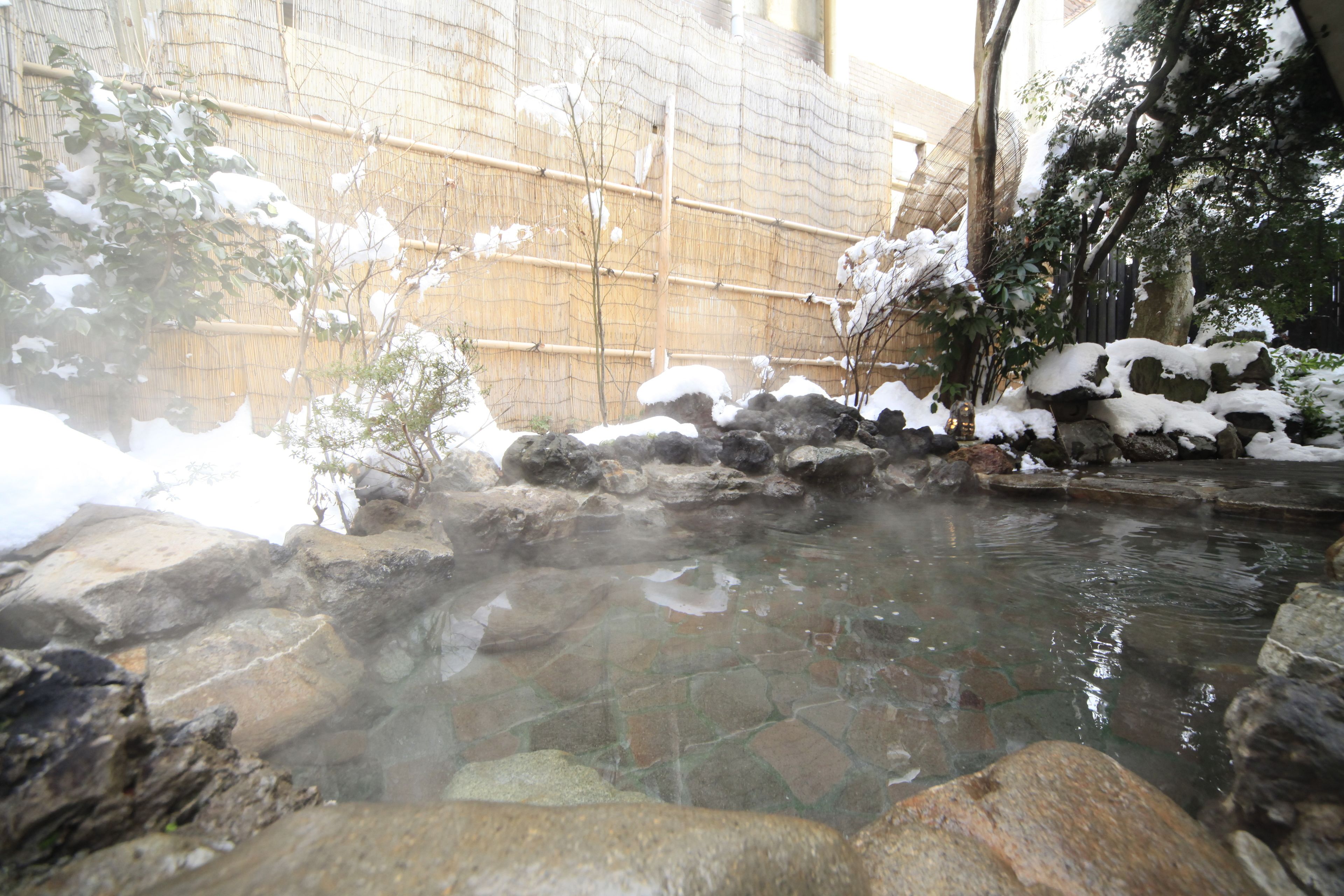 A hot spring area with stone steps leading into the water, surrounded by snow-covered rocks and bamboo fencing.