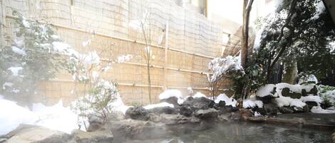A hot spring area with stone steps leading into the water, surrounded by snow-covered rocks and bamboo fencing.