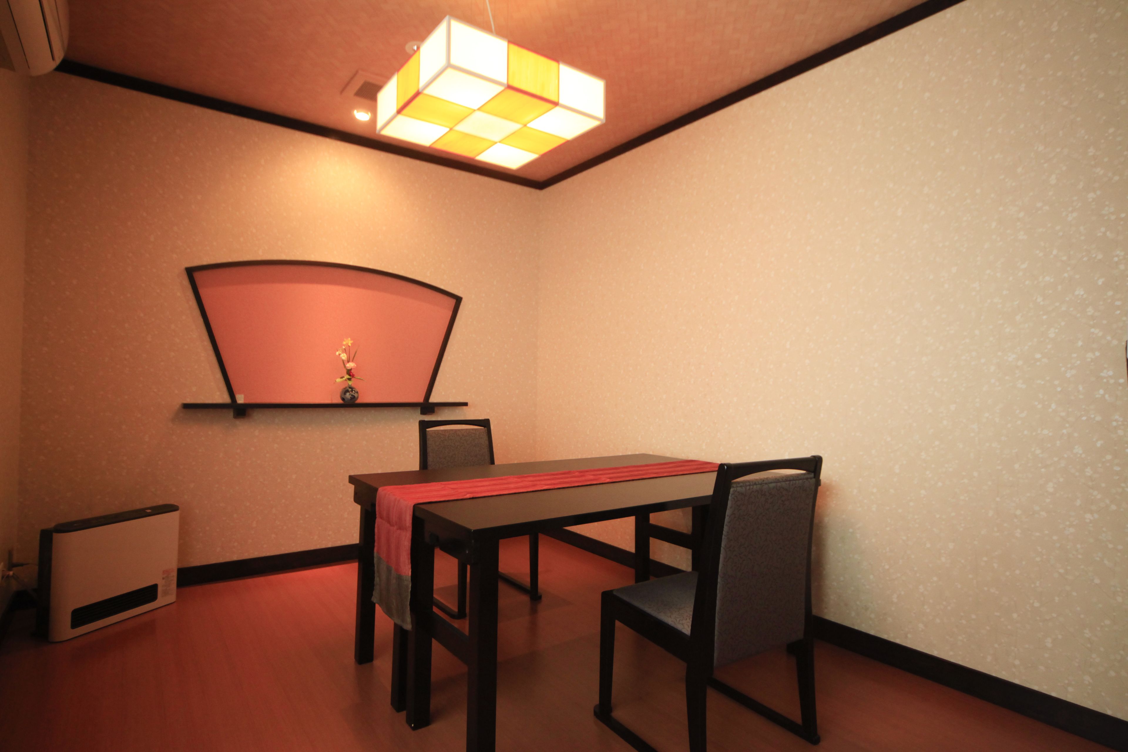 A room with a dining table, chairs, a red table runner, a wall-mounted shelf with a vase, and a ceiling light fixture.