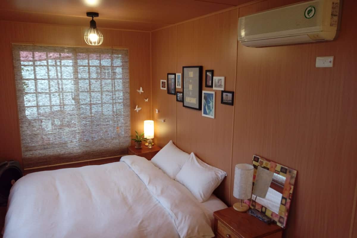 A neatly made bed with white linens, a bedside lamp, a wall-mounted air conditioner, and a framed picture collection on the wall.