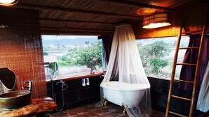 Une salle de bain rustique avec une baignoire placée sous une sorte de dais, un lavabo en bois et une vue sur l’extérieur.