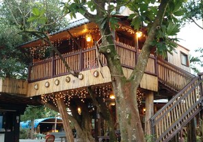 Una casa en el árbol con revestimiento de madera, un balcón con barandillas y luces de cuerda.