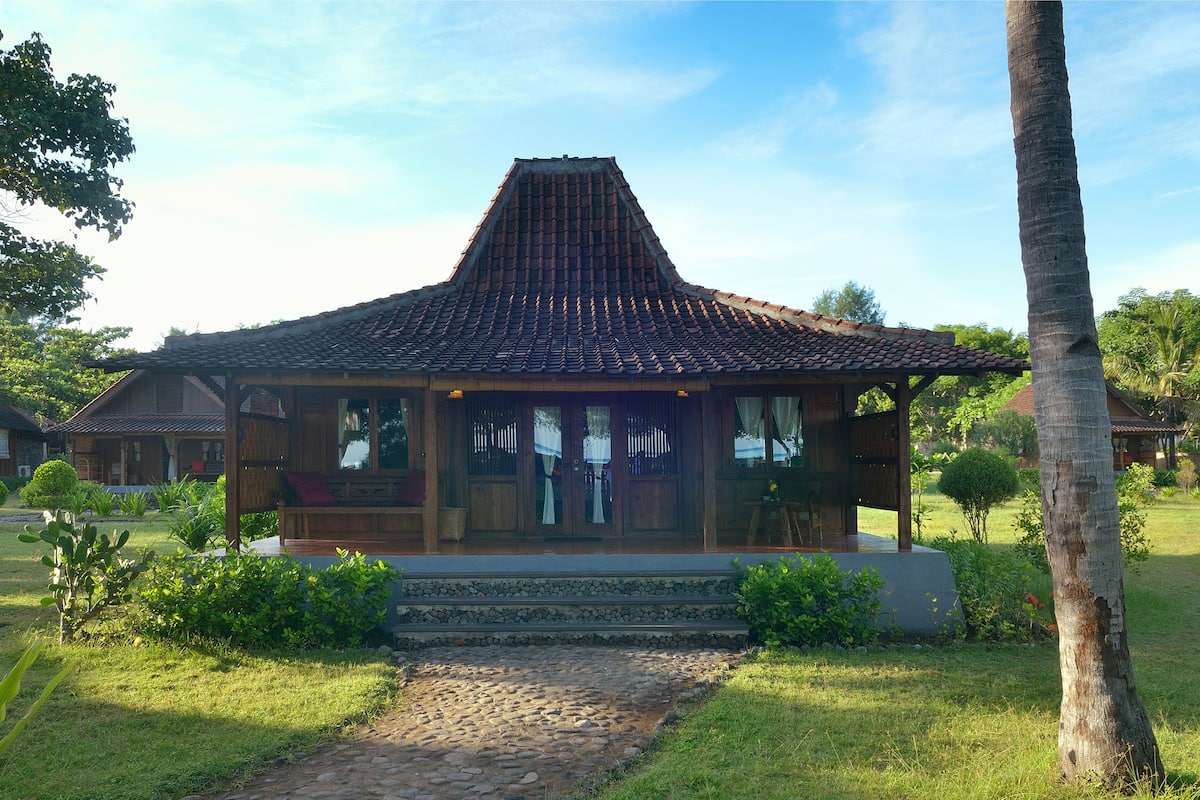 A traditional wooden house with a tiled roof, surrounded by lush greenery and a stone pathway.