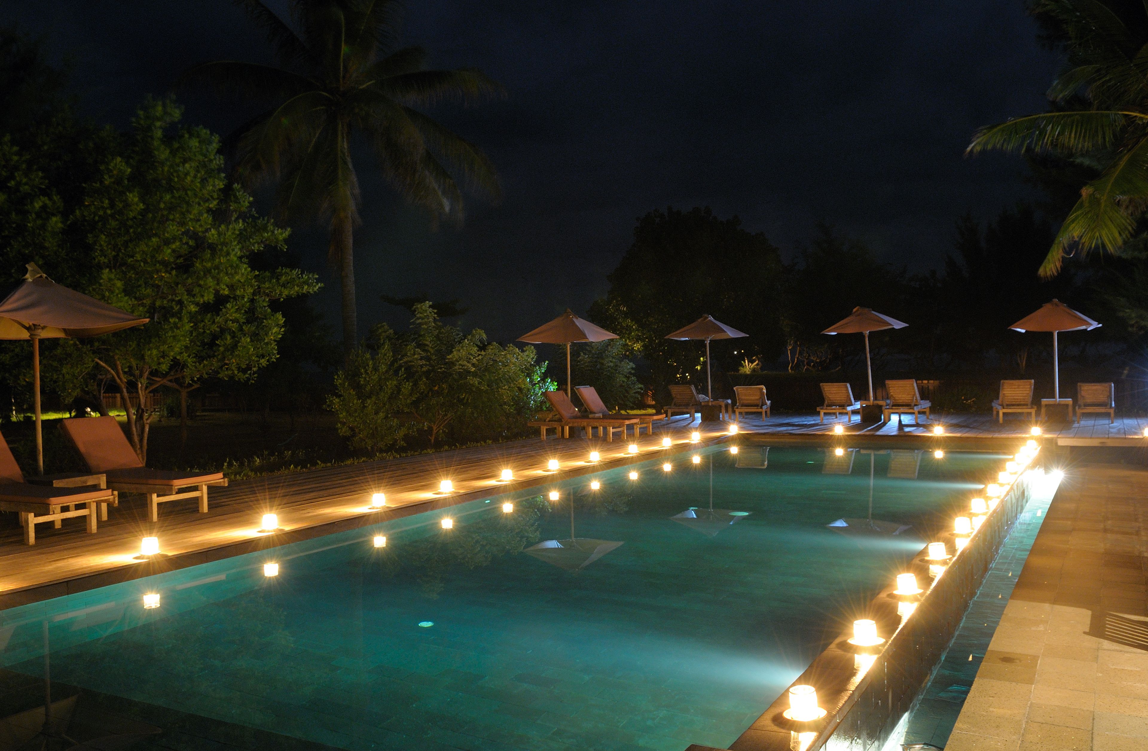 A nighttime pool area with lounge chairs and umbrellas, surrounded by palm trees.