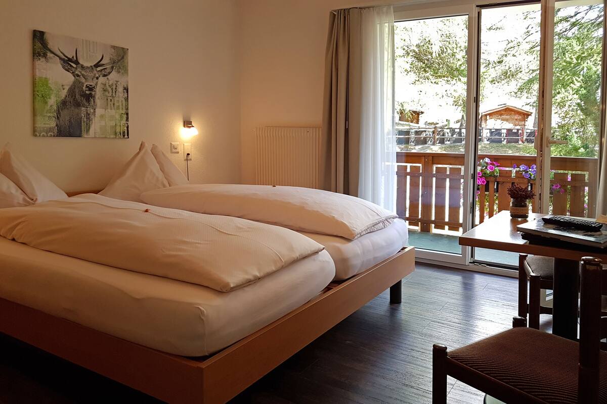 A double bed with white linens, a bedside table with a lamp, and a view of a balcony with flowers.