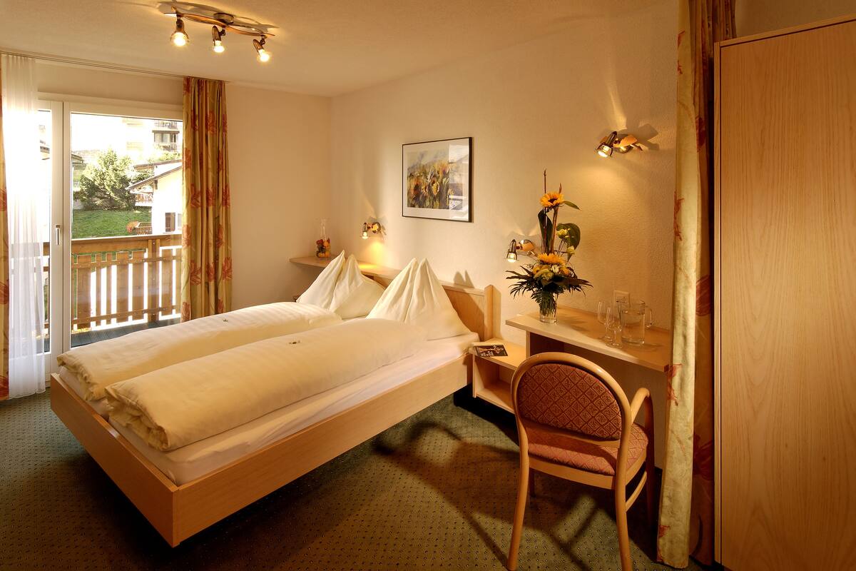 A hotel room with a bed, a desk, a chair, a window with curtains, a painting on the wall, and a balcony with a railing.