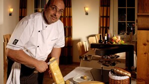 A chef preparing food in a well-lit kitchen with a dining table set for a meal, a wine bottle, and a basket of bread. - Parkhotel Saas- Fee (Saas-Fee)