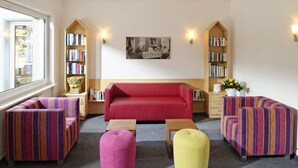 A cozy living room with a red sofa, striped armchairs, and ottomans. There are bookshelves on either side of the room, a coffee table, and a framed picture on the wall. - Parkhotel Saas- Fee (Saas-Fee)