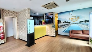 Un espace d’accueil d’hôtel comprenant un comptoir de réception, un écran numérique, un extincteur et une fresque murale représentant une piscine et une plage.