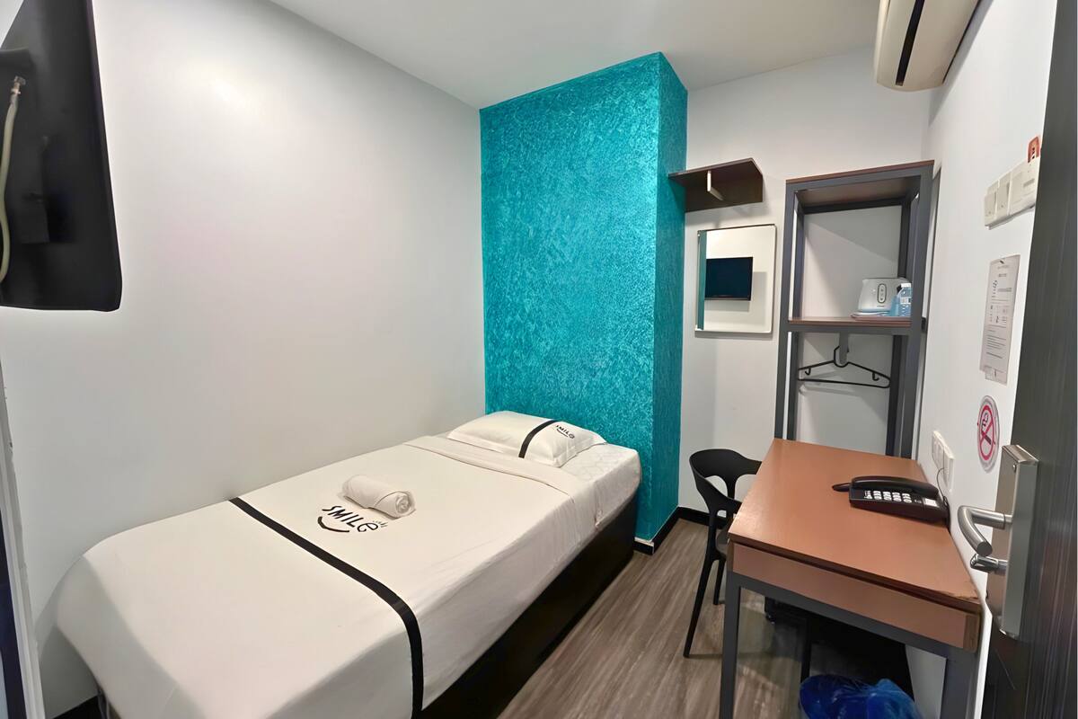Een kleine kamer met een enkel bed, een bureau, een stoel en een tv aan de muur.