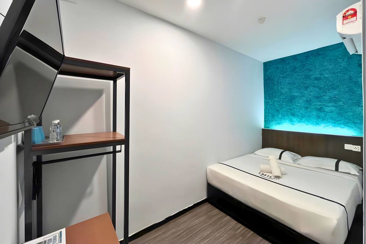 Hotelkamer met een bed, een klein tafeltje met een fles en een doos zakdoeken, een wandplank en een muur met een blauw textuurontwerp.