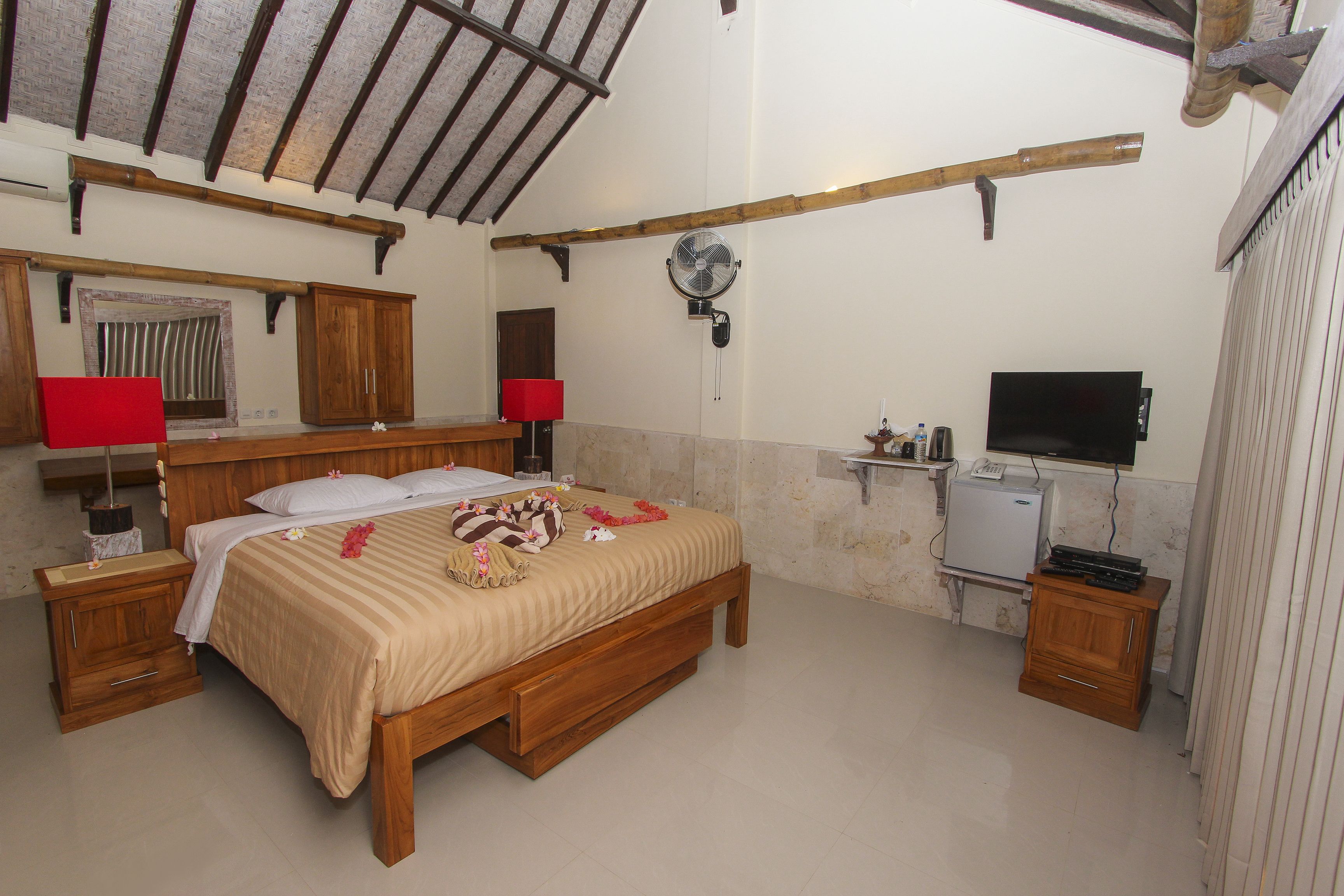 A bedroom with a wooden bed, a bedside table, a red lamp, a fan, a TV, and a small refrigerator.