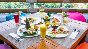 A table set with various dishes, drinks, and condiments, viewed from a balcony with a view of greenery. - Rinjani Lodge (Bayan)