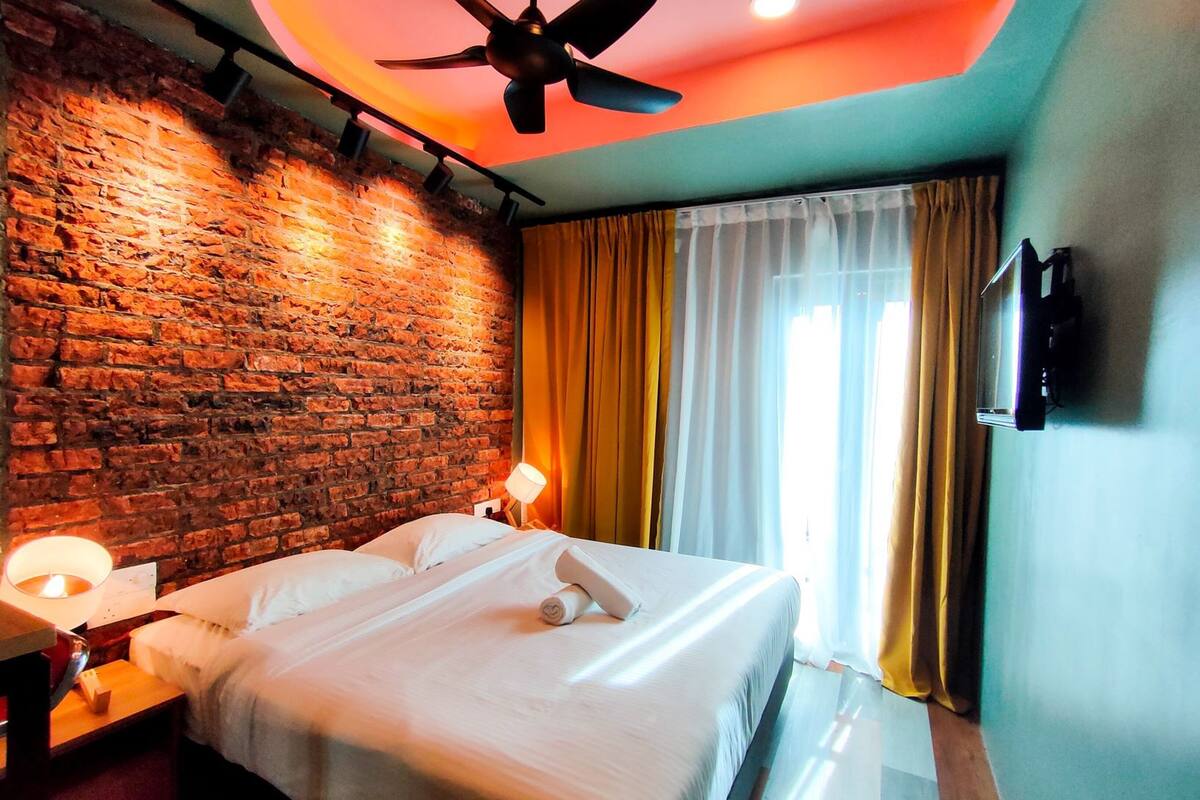 Hotelkamer met een bakstenen muur, een bed met witte lakens, een plafondventilator en een aan de muur bevestigde televisie.