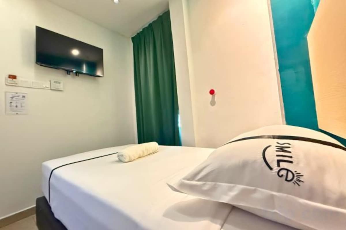 Een hotelkamer met een bed, een flatscreen televisie aan de muur, een groen gordijn en een kussen met een vrolijk gezicht.