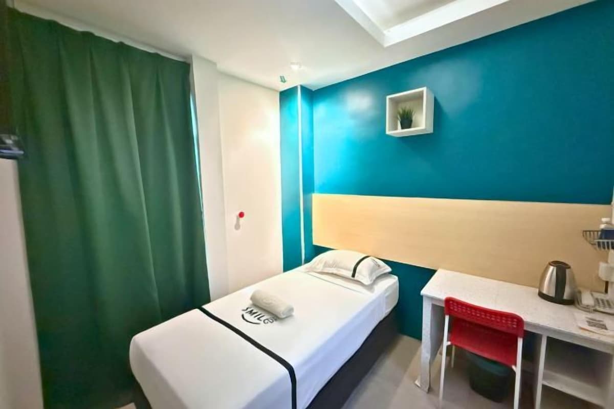 Ein Einzelbett mit weißem Kopfteil, ein kleiner Tisch und ein roter Stuhl, sowie eine Wand in Blau.