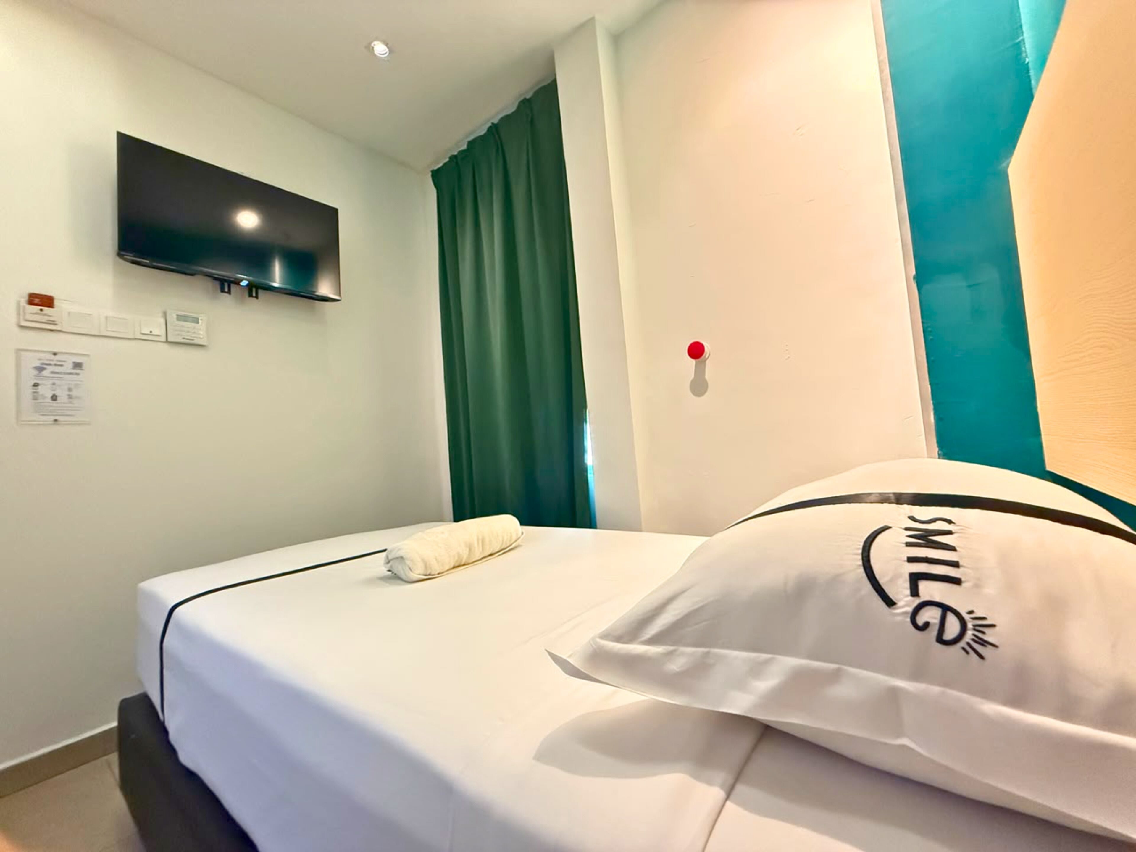 A hotel room with a bed, a flat-screen TV mounted on the wall, a red emergency button, and a teal-colored wall.