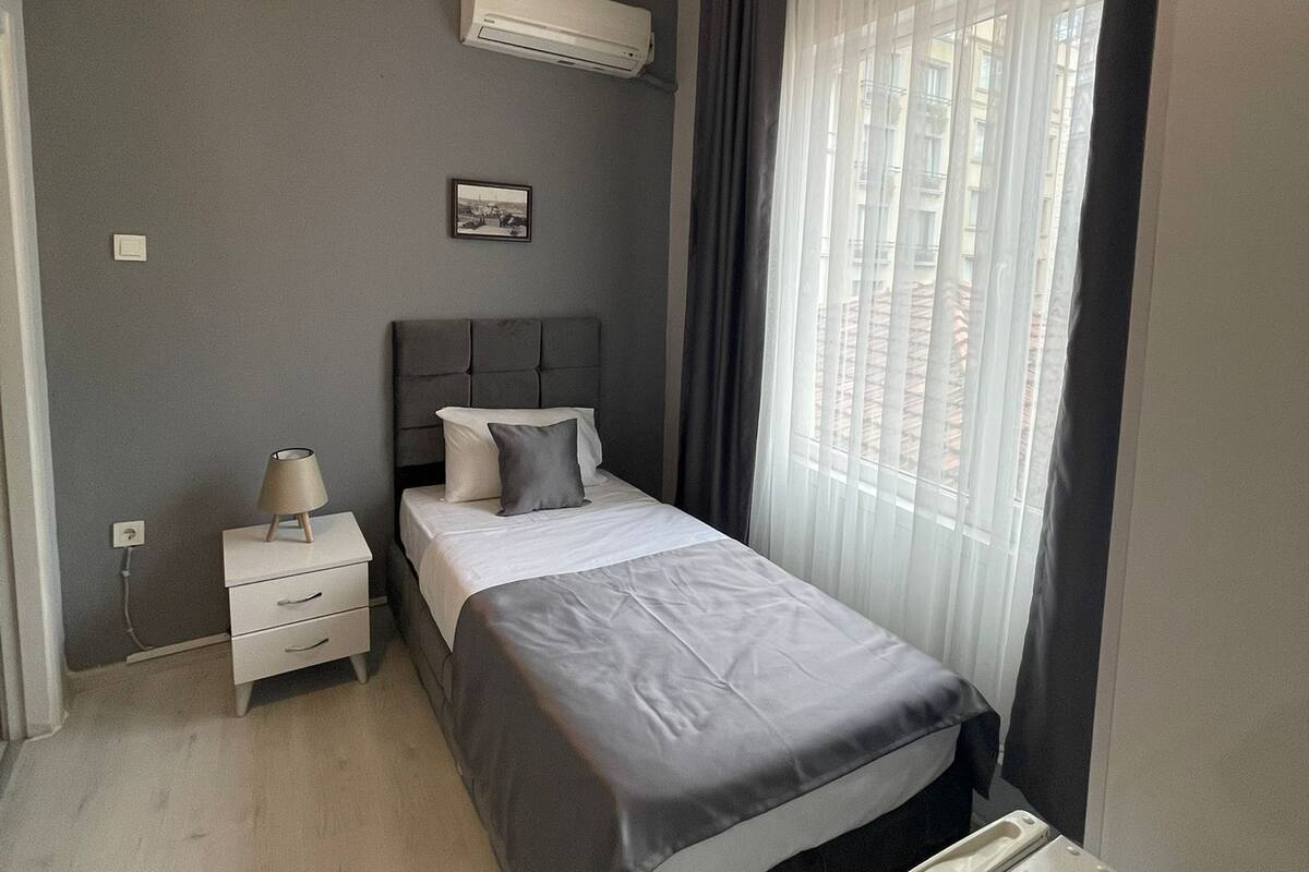 A bedroom with a bed, bedside table, lamp, air conditioning unit, and a window with curtains.