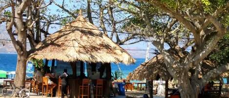 A beachside area with thatched-roof huts, a bicycle, and a hammock.