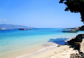 A beach with clear turquoise water, boats, and rocky shore.