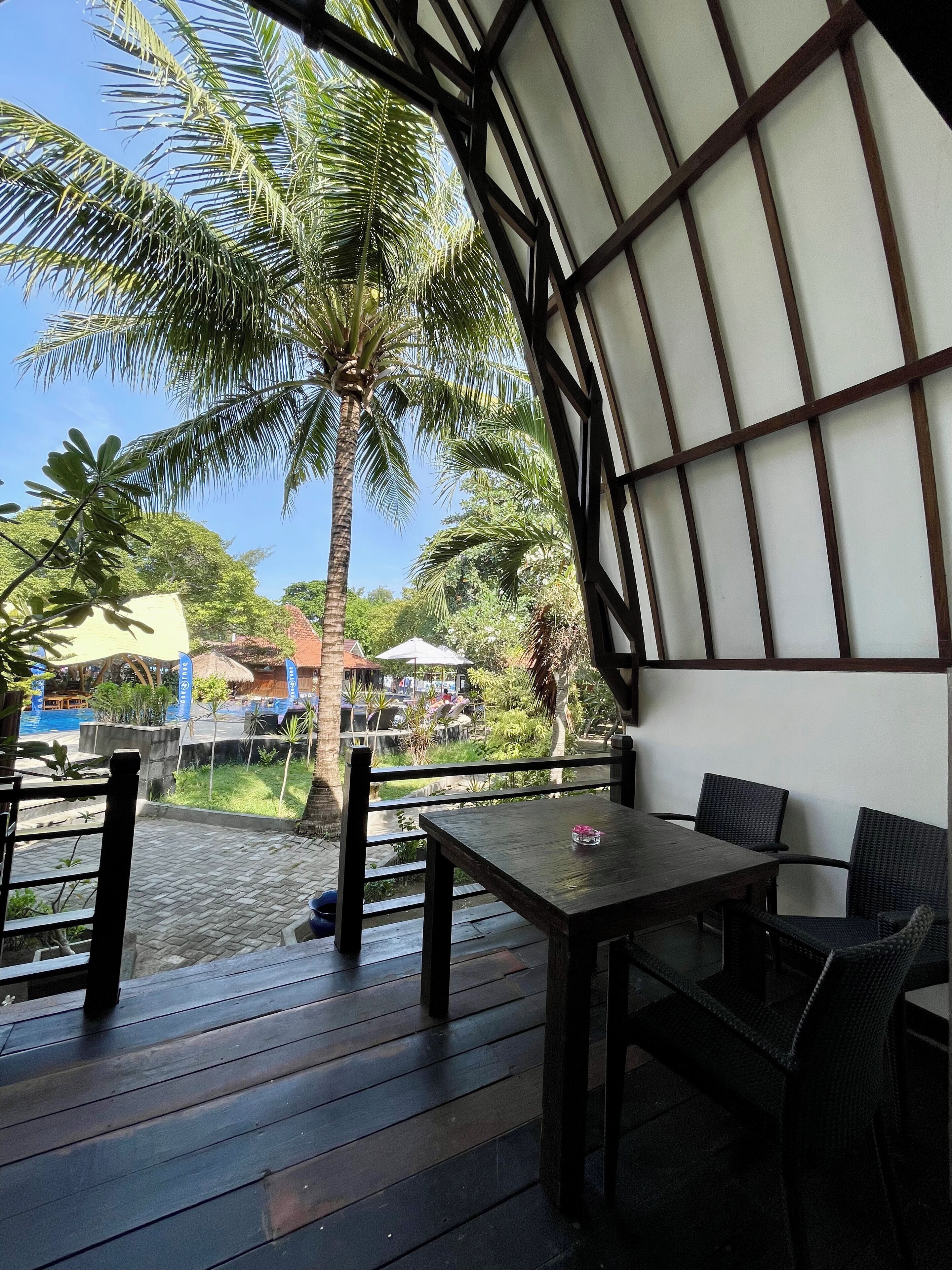 A covered outdoor seating area with a table and chairs, overlooking a water body and palm trees.