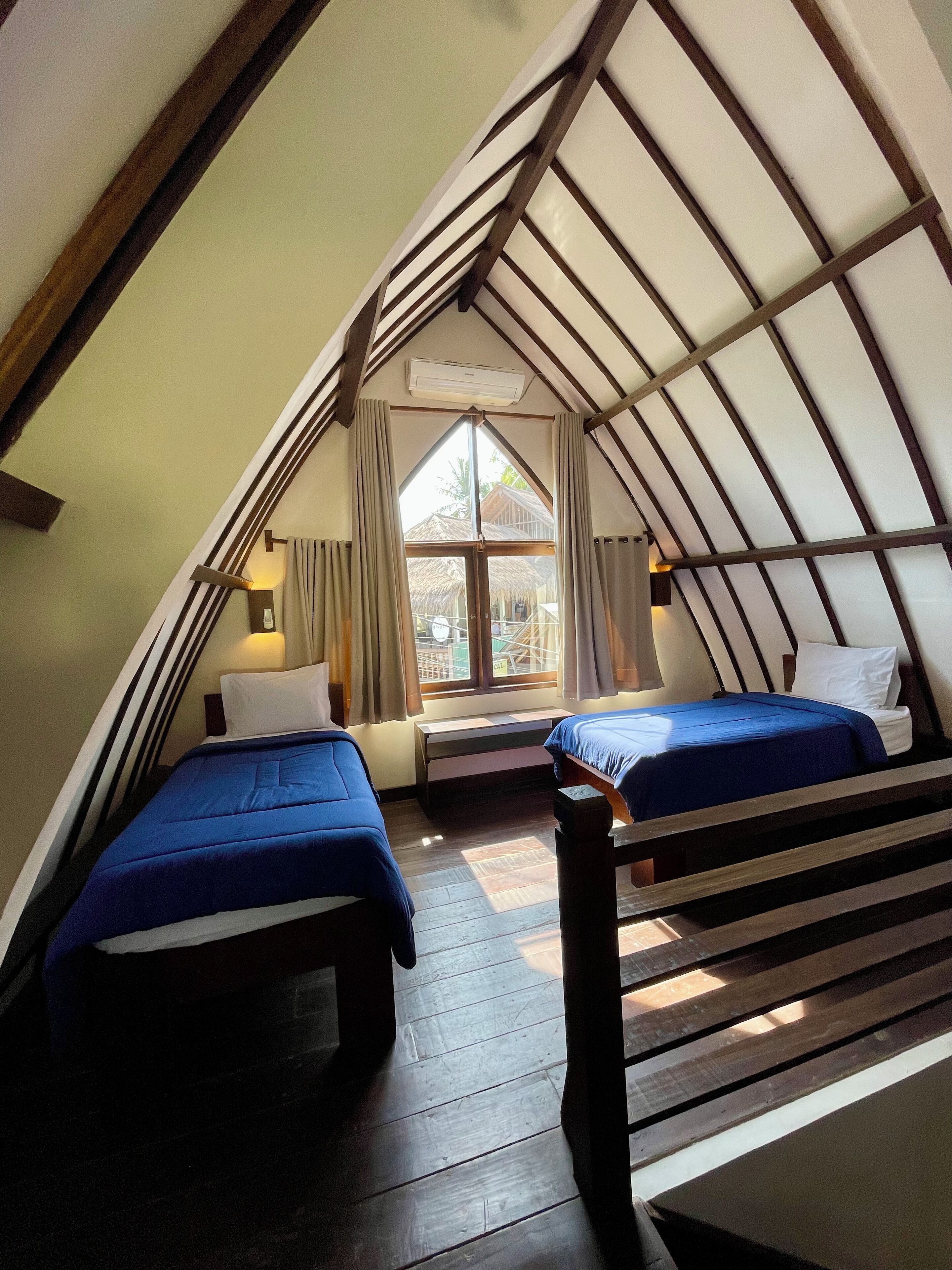 A cozy attic bedroom with two beds, wooden beams, a large window, and a view of the outside.
