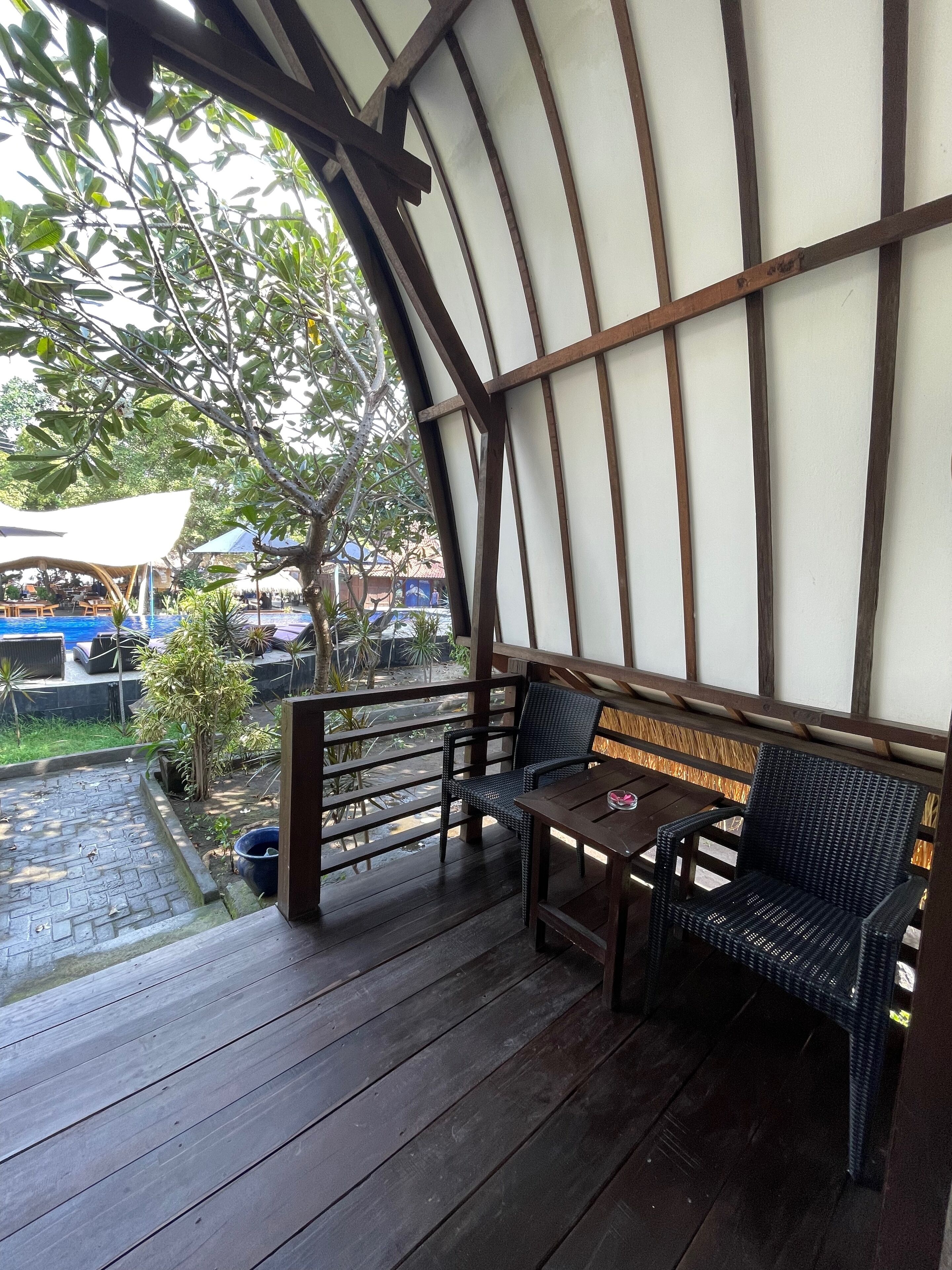 A covered outdoor seating area with a wooden bench and table, surrounded by a wooden railing and a view of a pool and greenery.