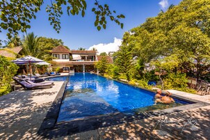 A swimming pool with lounge chairs and umbrellas, surrounded by lush greenery and a house with a tiled roof.