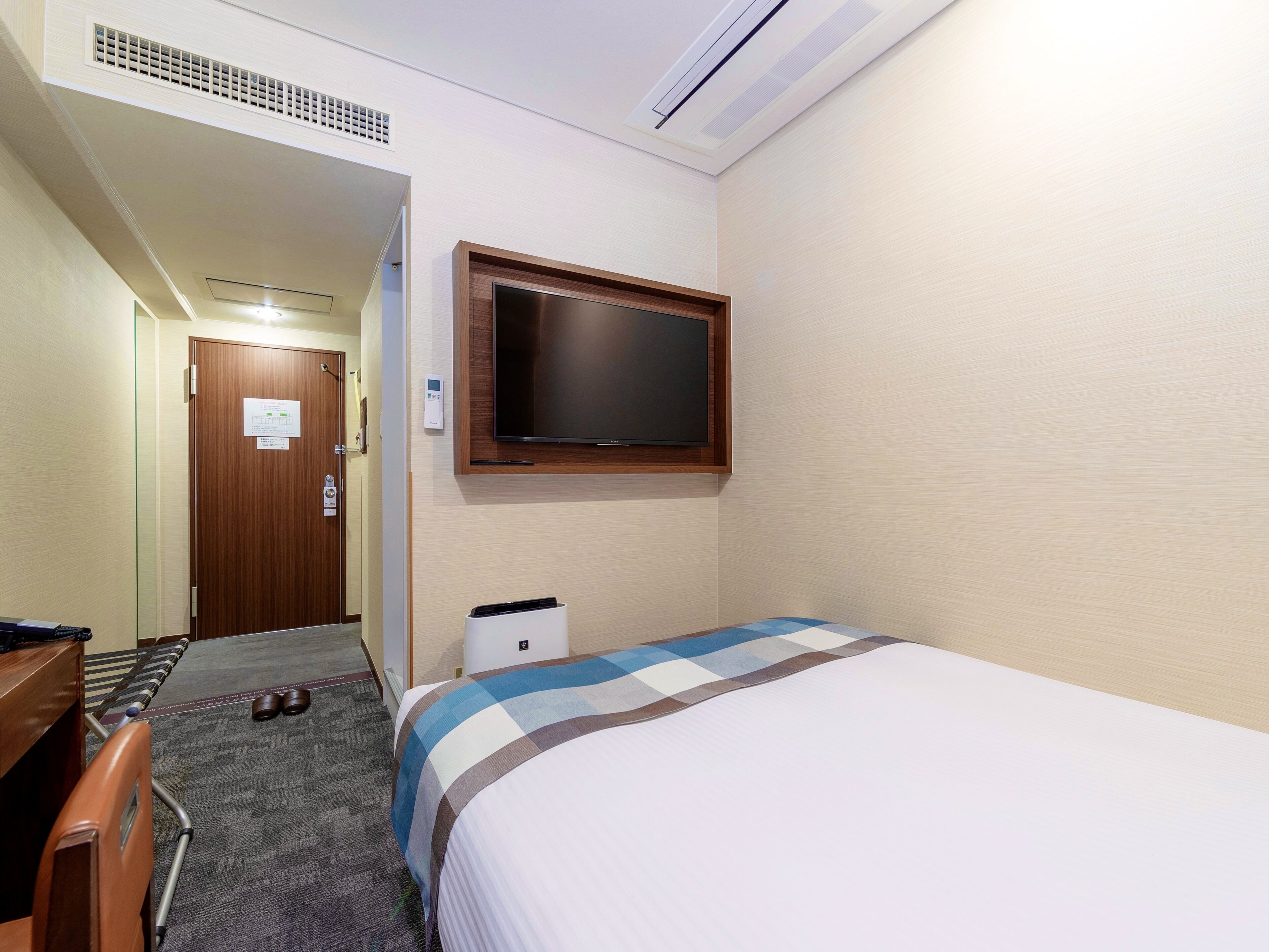 A hotel room with a bed, a television mounted on the wall, a desk with a chair, and a small bedside table.
