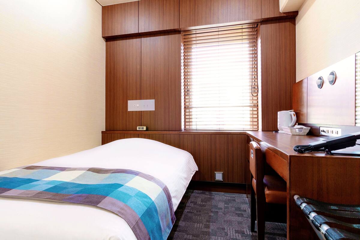 A hotel room with a bed, a desk, a chair, a window with blinds, a thermostat, and a telephone.