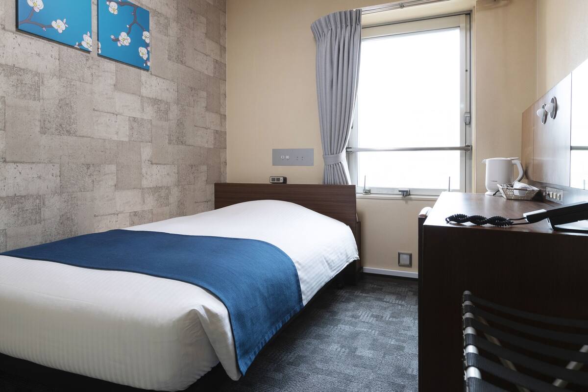 A hotel room with a bed, a desk, a chair, a window with curtains, and a wall with a brick pattern.