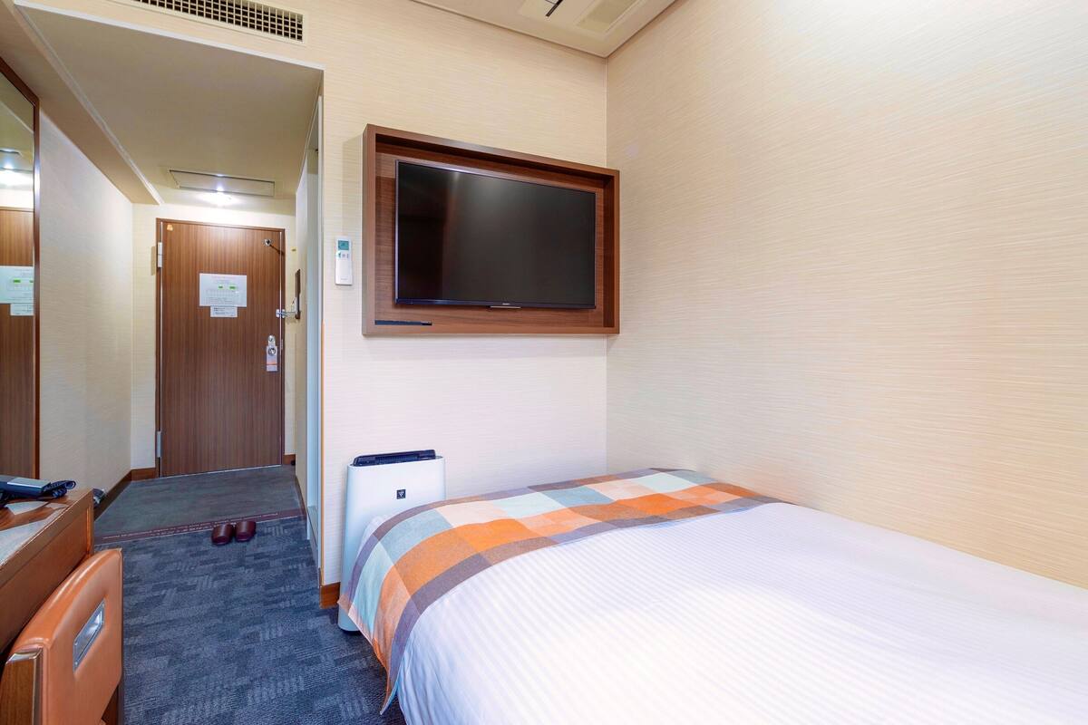 A hotel room with a bed, a desk, a chair, a television mounted on the wall, and a door leading to another room.