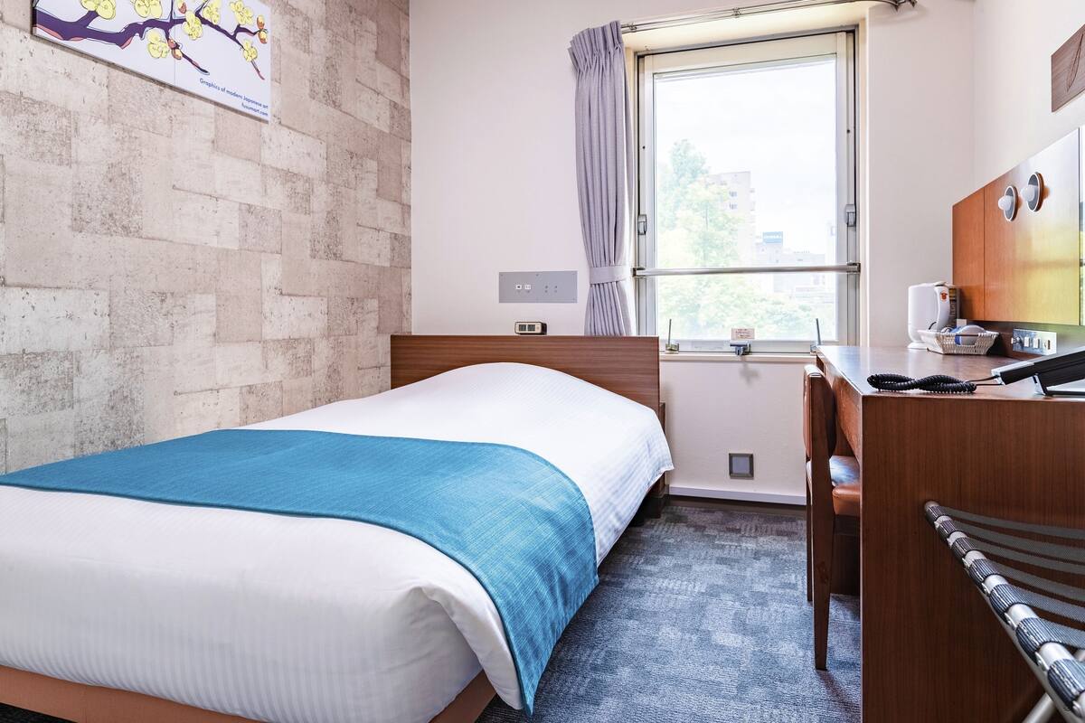 A hotel room with a bed, a desk, a chair, a window with curtains, and a wall-mounted TV.