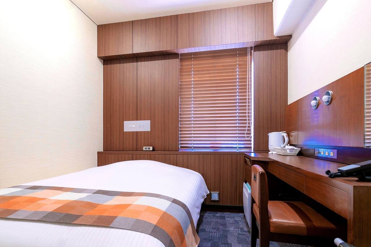 A hotel room with a bed, a desk, a chair, a window with blinds, and a control panel on the wall.
