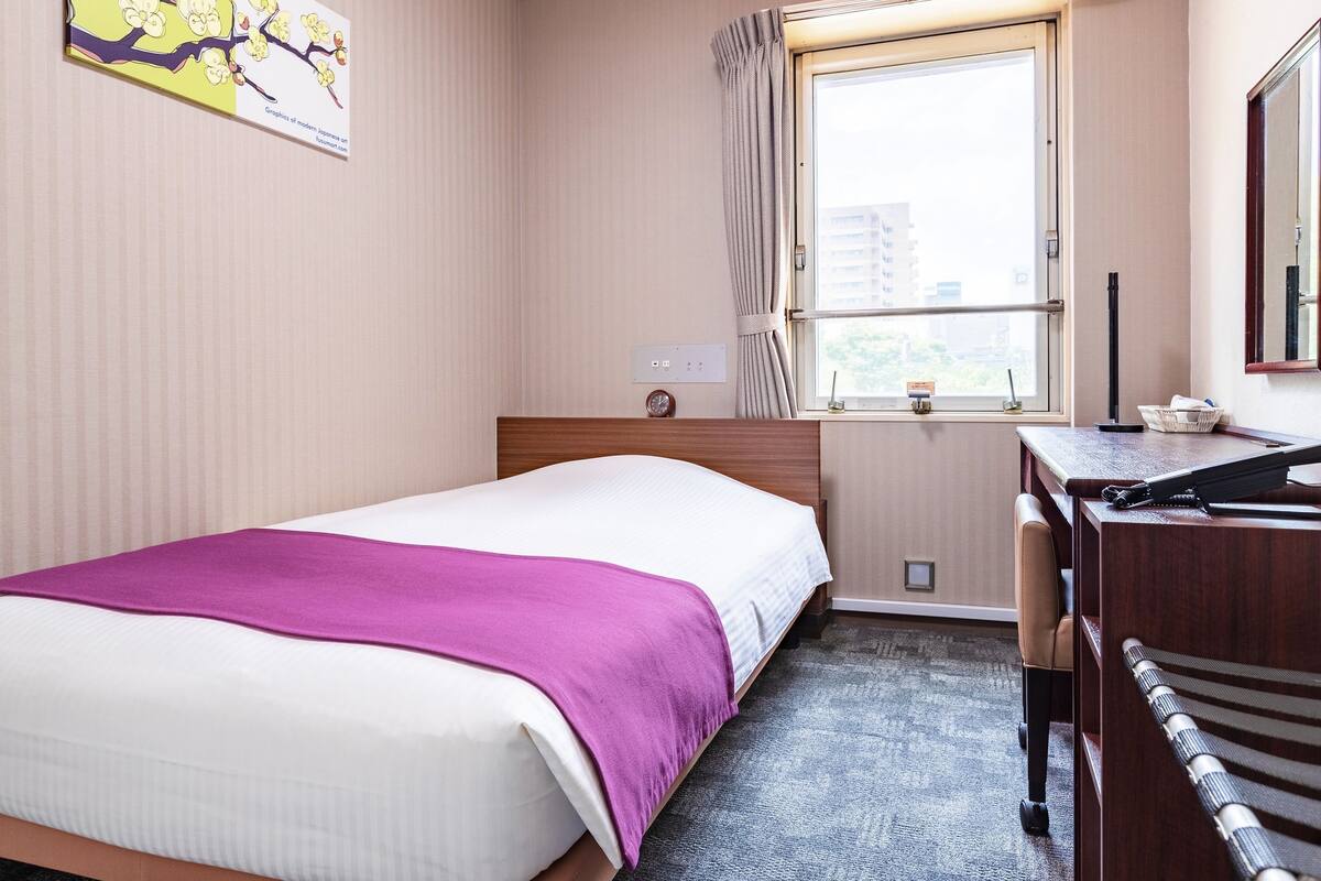 A hotel room with a bed, a desk, a chair, a window with curtains, a wall-mounted clock, and a painting.