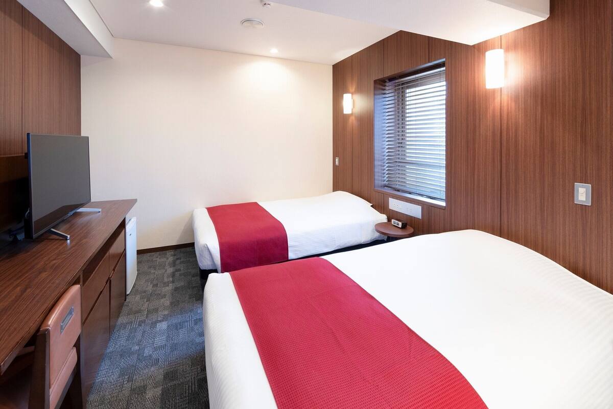 A hotel room with two beds, a desk, a television, and a window with blinds.