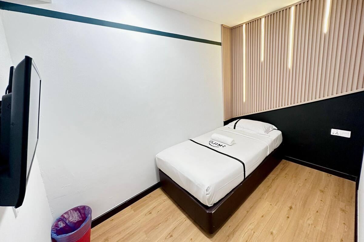 A small, modern hotel room with a single bed, a flat-screen TV mounted on the wall, and a wooden floor.