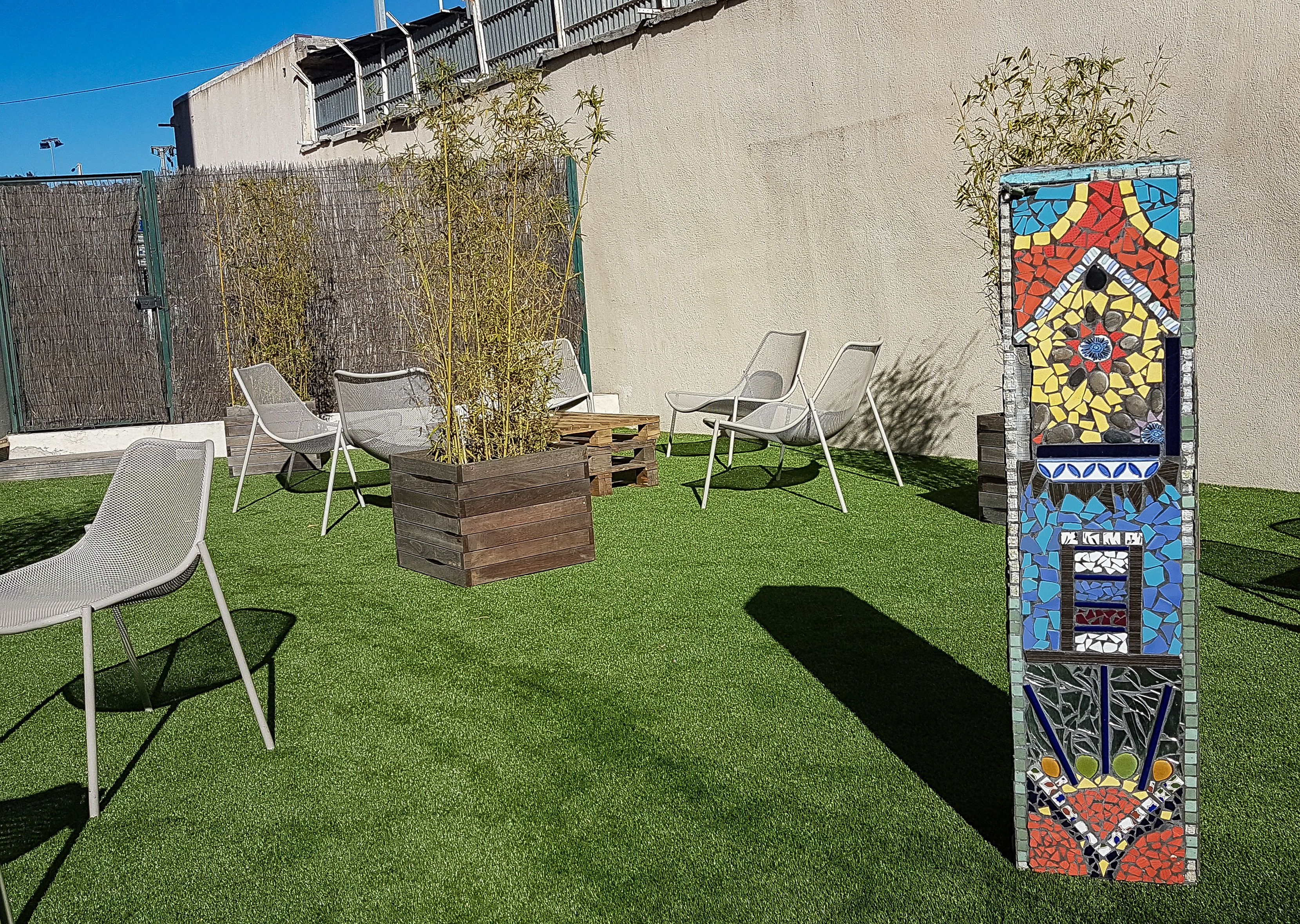 Un espace patio avec un revêtement de sol en gazon synthétique, un bac à fleurs en bois contenant des plantes et deux chaises blanches en tissu à mailles.