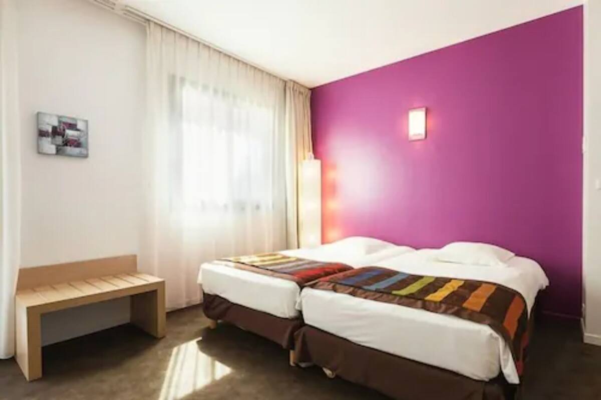 Habitación de hotel con dos camas, un banco, una ventana con cortinas y una pared morada.