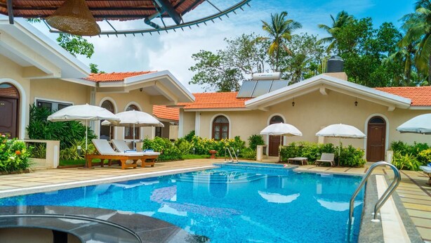 A swimming pool with lounge chairs and umbrellas, surrounded by greenery and a building with a tiled roof. - Jacks Resort (Vagator)