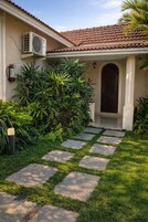 A house with a tiled roof, a stone pathway leading to a dark entrance, and lush greenery around.
