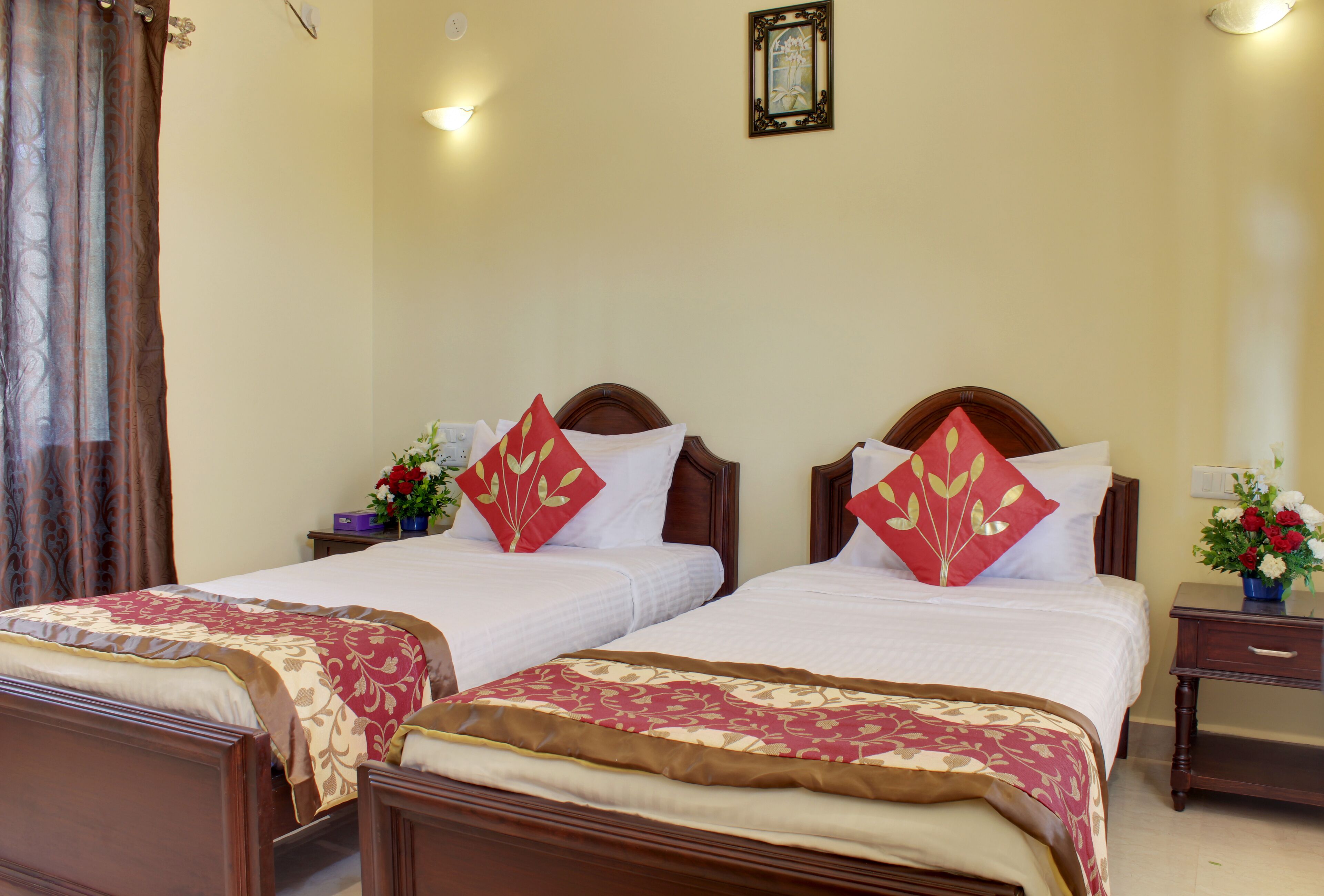 A hotel room with two single beds, each with a wooden headboard, white bedding, and red decorative pillows. There is a nightstand with a vase of flowers and a small table lamp on the right side.