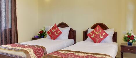 Habitación de hotel con dos camas individuales, cada una con cabecero de madera, ropa blanca y almohadas decorativas rojas. A la derecha, un mesita de noche con un jarrón de flores y una lámpara de mesa.