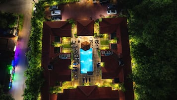 Vista aérea de una propiedad con piscina rodeada de sillas de descanso y sombrillas, un patio cubierto y una calle con coches estacionados.